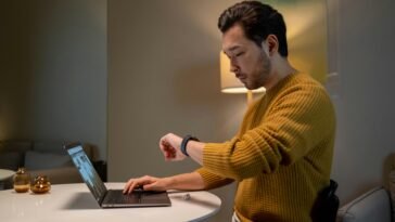 Man in Brown Sweater Looking at Time on His Smartwatch while Using Laptop